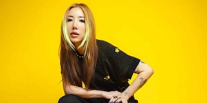 Person with long light brown hair and green streaks, wearing a black outfit, sits against a bright yellow background.