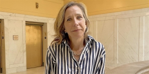 A woman wearing a blue and white striped shirt stands indoors in a marble hallway with elevators in the background.