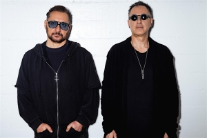 Two men wearing dark sunglasses and black clothing stand against a plain white wall, looking straight at the camera.