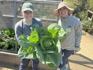 Two people in hats and casual clothes stand in a garden, smiling and holding a large cabbage.