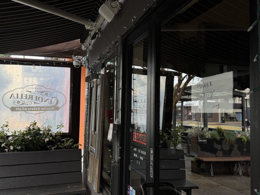 Exterior view of a bakery café with a "Closed" sign on the glass door and plants on the patio; reflections and handwritten signs are visible in the window.