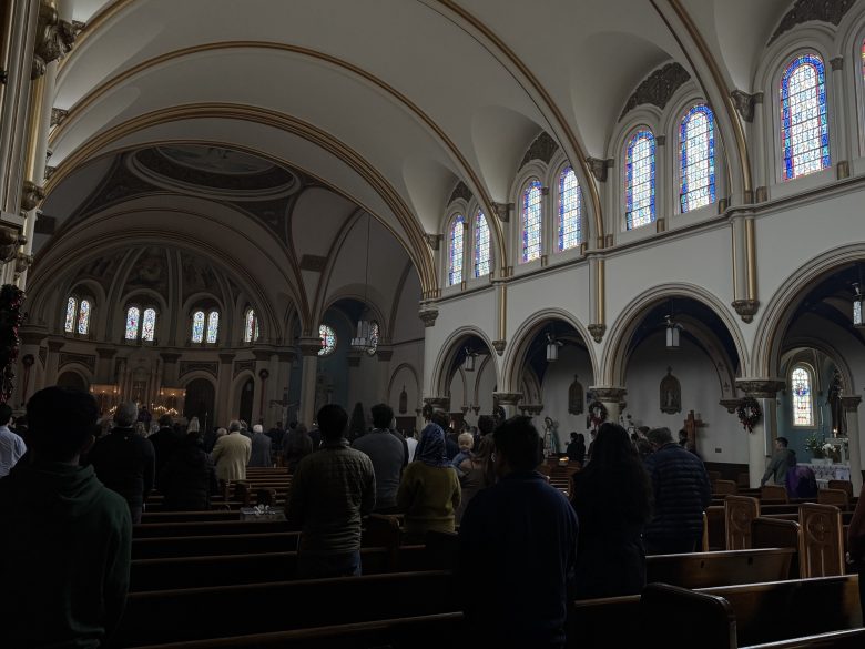 People attend a service inside a church with arched ceilings and stained-glass windows; the congregation is standing in pews facing the altar.