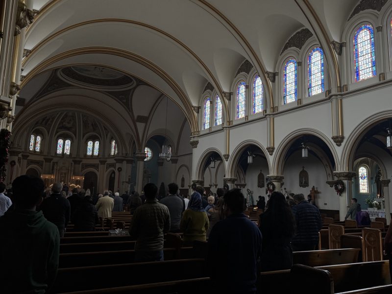 People attend a service inside a church with arched ceilings and stained-glass windows; the congregation is standing in pews facing the altar.
