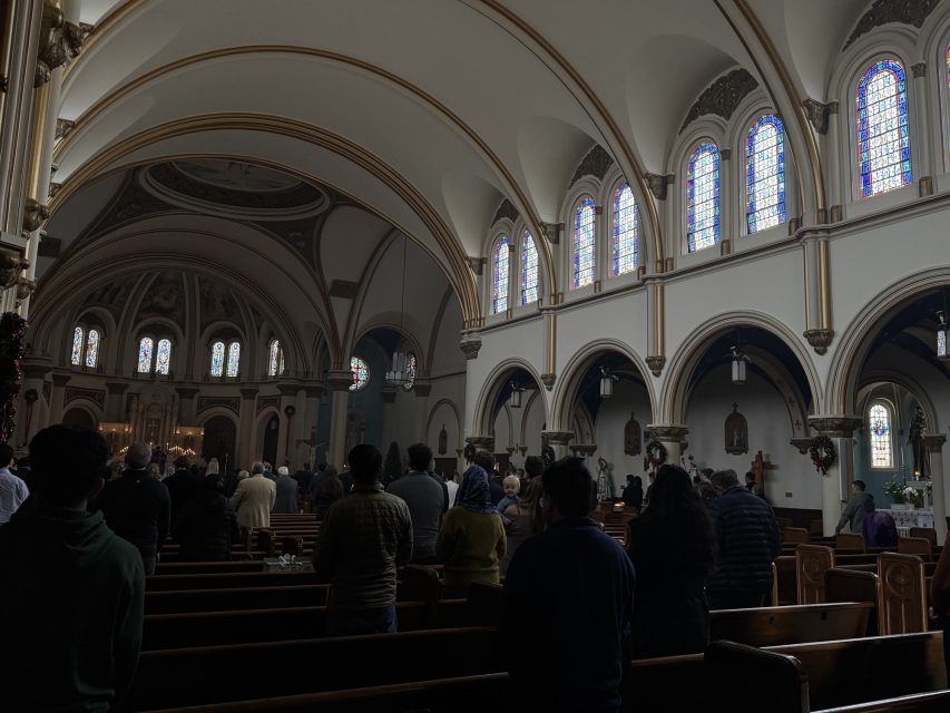 People attend a service inside a church with arched ceilings and stained-glass windows; the congregation is standing in pews facing the altar.