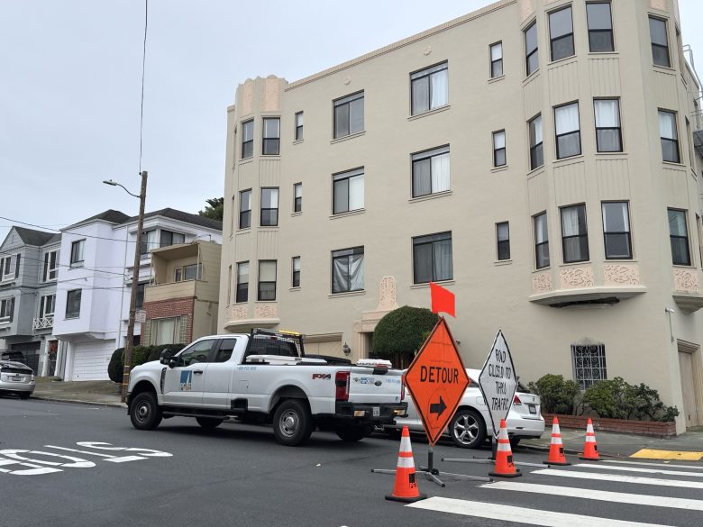 A white utility truck is parked by an intersection with orange cones, a "Detour" sign, and "Road Closed to Thru Traffic" sign in front of an apartment building.