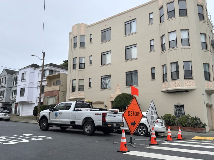 A white utility truck is parked by an intersection with orange cones, a "Detour" sign, and "Road Closed to Thru Traffic" sign in front of an apartment building.