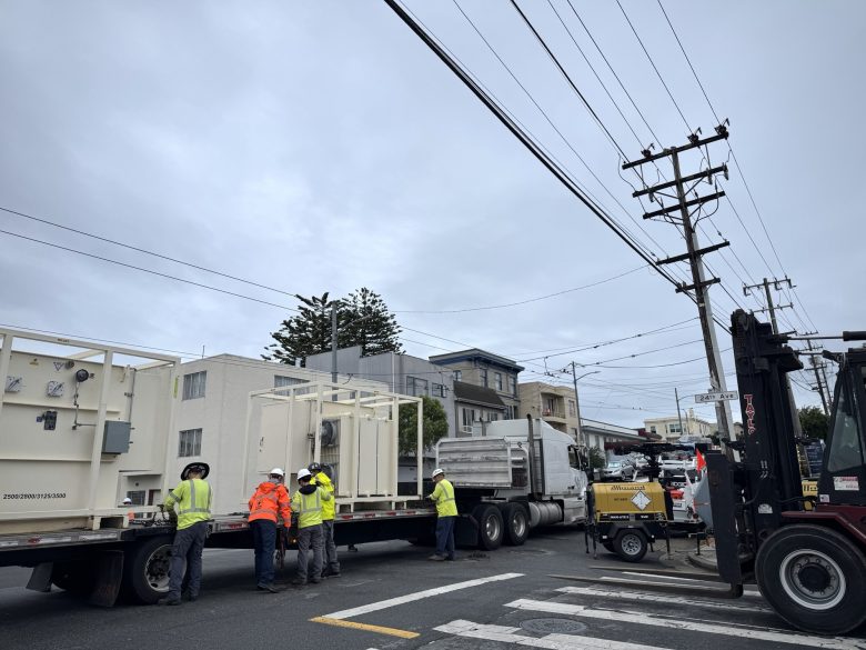 Several construction workers in safety vests stand by a flatbed truck loaded with large structures on a city street, with a forklift and utility lines overhead.