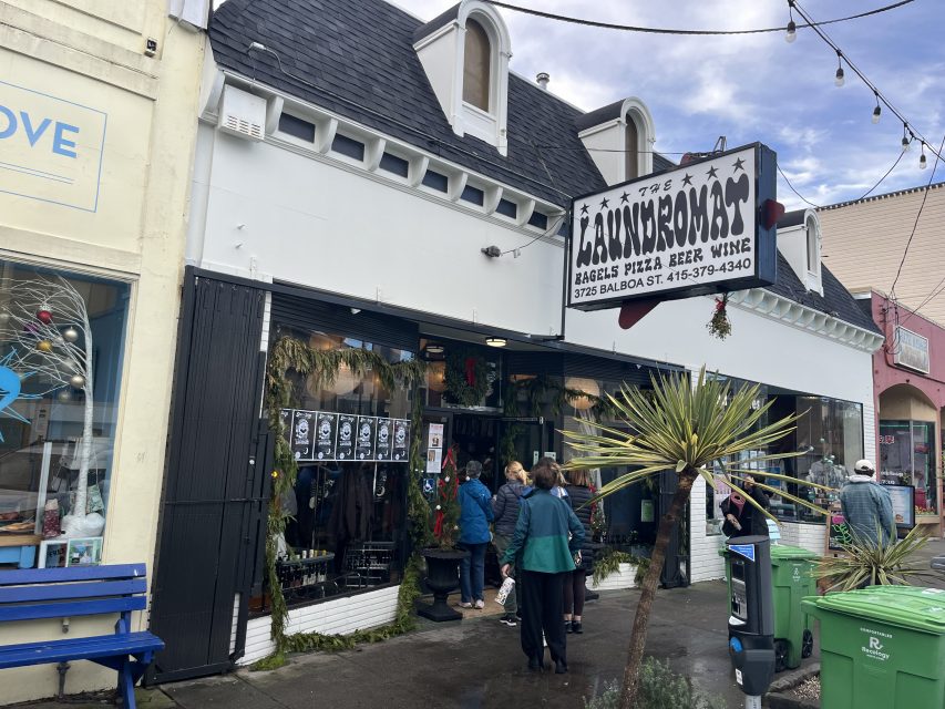 People stand outside The Laundromat, a shop offering bagels, pizza, beer, and wine on Balboa Street. The storefront is decorated with greenery and has a black and white sign.