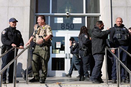 Several uniformed law enforcement officers, including Derrick Lew, stand and talk outside the entrance of a government building marked "850.