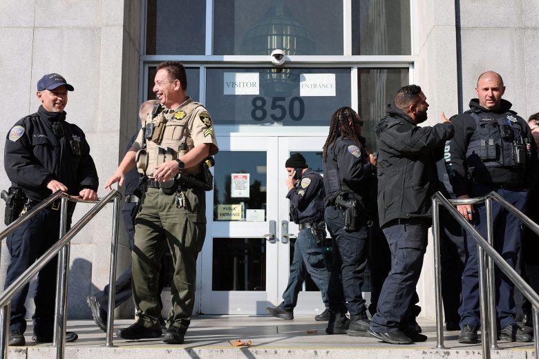 Several uniformed law enforcement officers, including Derrick Lew, stand and talk outside the entrance of a government building marked "850.