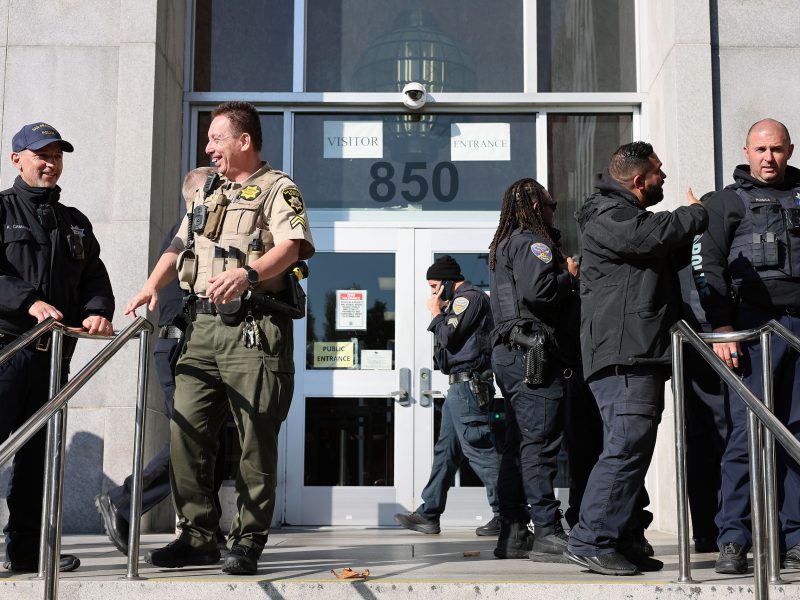 Several uniformed law enforcement officers, including Derrick Lew, stand and talk outside the entrance of a government building marked "850.