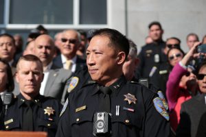 Police officer Derrick Lew, in uniform, stands at a podium outdoors, surrounded by fellow officers and people, with a building in the background.