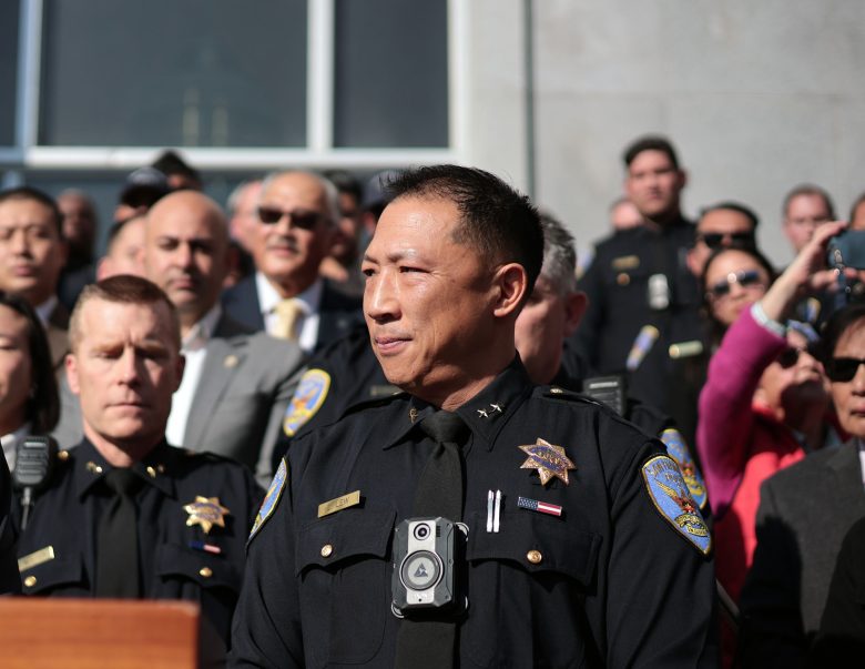 Police officer Derrick Lew, in uniform, stands at a podium outdoors, surrounded by fellow officers and people, with a building in the background.