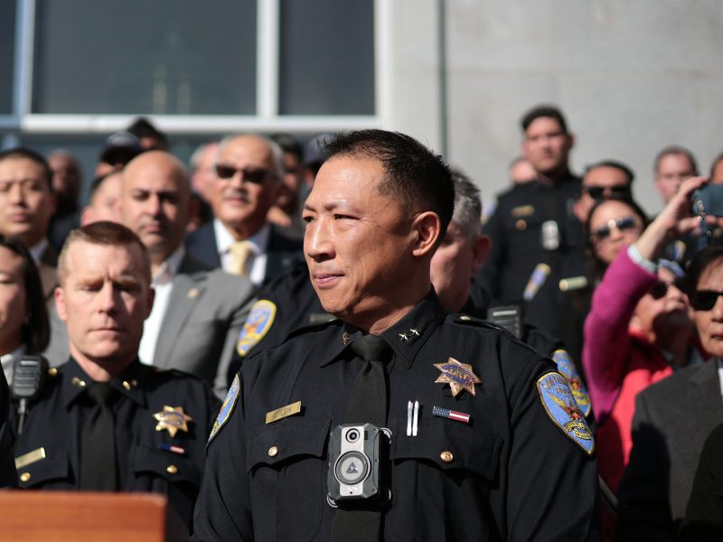 Police officer Derrick Lew, in uniform, stands at a podium outdoors, surrounded by fellow officers and people, with a building in the background.