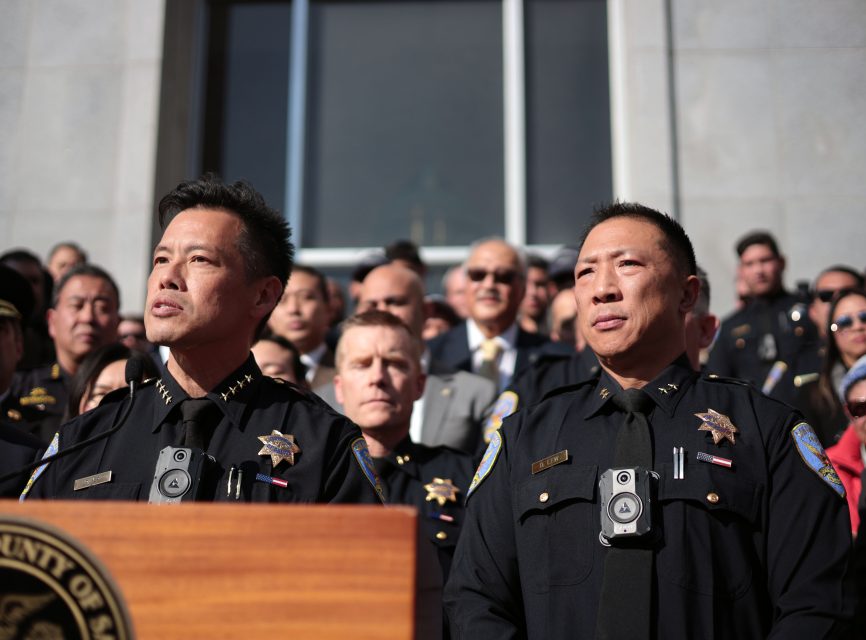 Two uniformed police officers stand at a podium outdoors, with Derrick Lew and a crowd of people in formal attire gathered behind them.