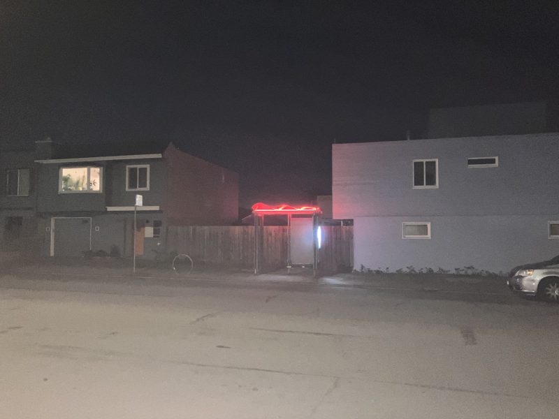 A dark street at night with two houses; a red neon light glows above a fenced gate between the buildings. A parked car and a bicycle are visible.