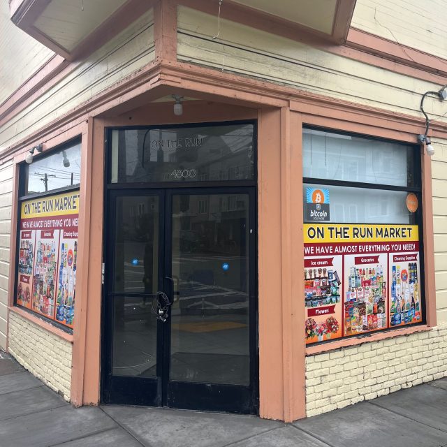 A corner storefront with large windows displays “On The Run Market” signs advertising groceries and a Bitcoin sticker on the door. The store appears closed.