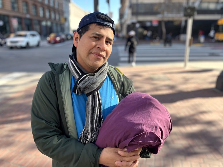 A man wearing a cap, scarf, and jacket stands on a city sidewalk holding a rolled-up purple blanket, with buildings and cars in the background.