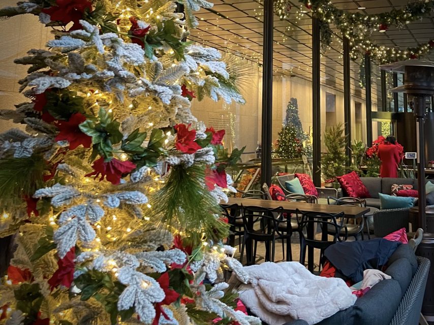 A snow-flocked Christmas tree with red poinsettias and lights stands next to a patio seating area decorated with Christmas garlands and ornaments.