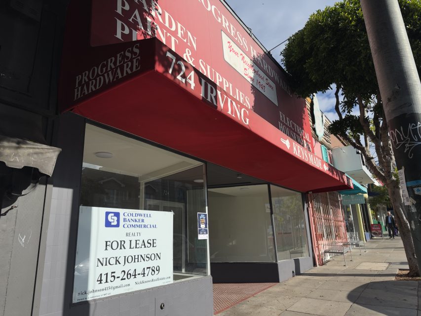 A vacant storefront with a "FOR LEASE" sign displayed in the window on a sunny urban street; the awning above reads "Progress Hardware.