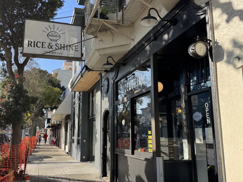Street view of the Rice & Shine restaurant entrance with an "OPEN" sign, sidewalk, and nearby shops visible under clear daylight.