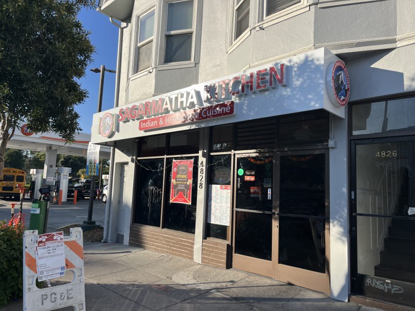Street view of Sagarmatha Kitchen, an Indian and Himalayan cuisine restaurant with a white and red sign, located next to a gas station and a school bus in the background.