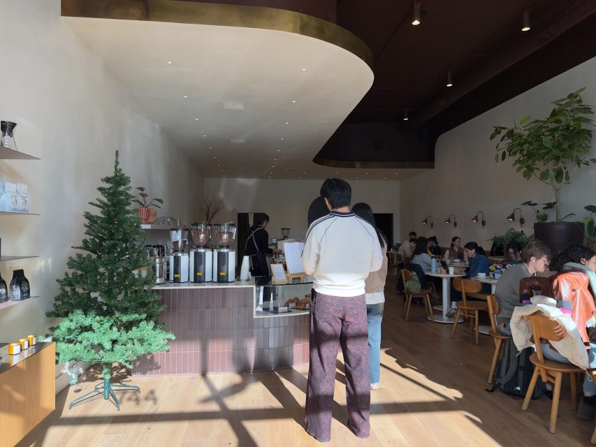 A man stands at a coffee shop counter near a small artificial Christmas tree, while several people sit at tables in the sunlit dining area.