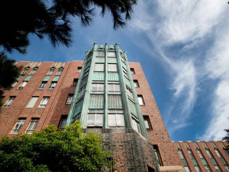 A brick building with a copper-green art deco facade rises into a blue sky with wispy clouds, framed by leafy trees.