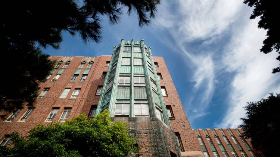 A brick building with a copper-green art deco facade rises into a blue sky with wispy clouds, framed by leafy trees.