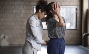 Two women stand close together indoors; one gently places her hand on the other's head and back, as if assisting with posture or movement.