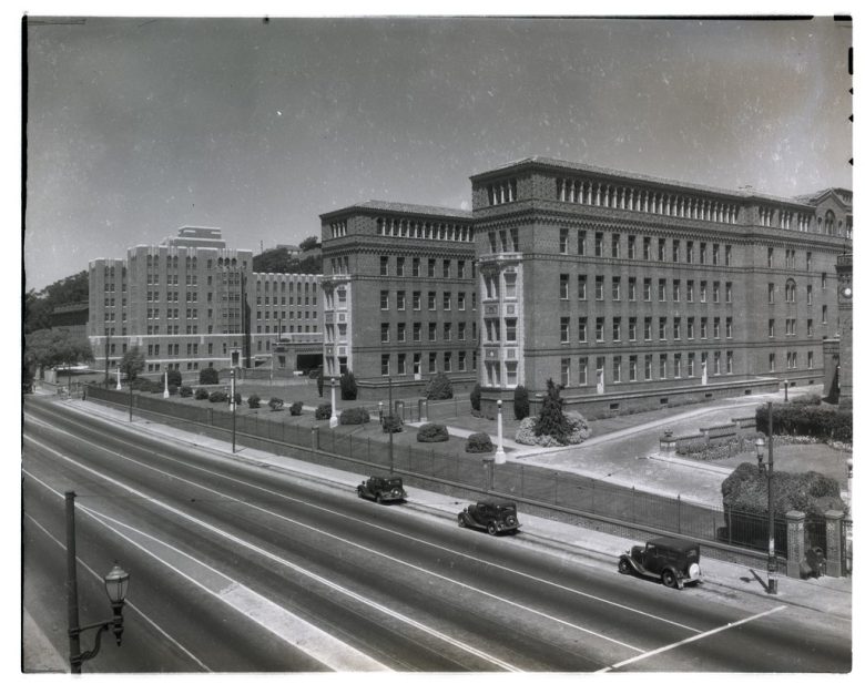 Black-and-white photo of large brick institutional buildings beside a wide, empty road with a few vintage cars parked along the curb.