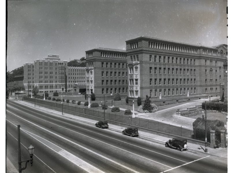 Black-and-white photo of large brick institutional buildings beside a wide, empty road with a few vintage cars parked along the curb.