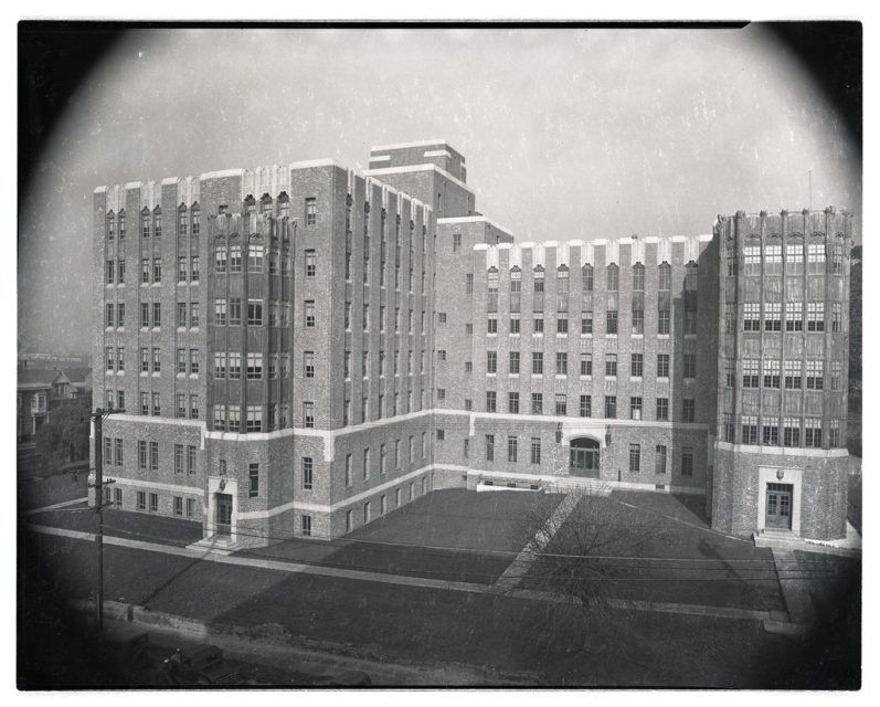 Black and white photo of a large, multi-story brick building with rectangular windows, flat rooflines, and an empty lawn in front.