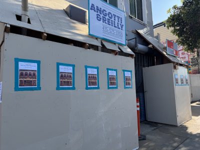 Construction site with a wooden barrier displaying multiple identical posters taped in blue; an "Ancotti Realty" sign is visible above the barrier.