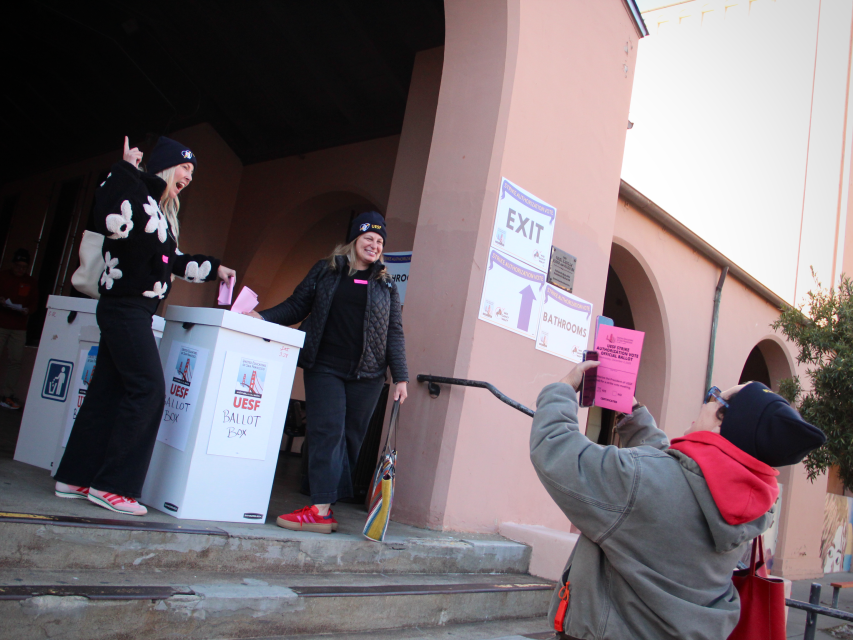 Three people stand outside a building near ballot boxes; one person poses with papers while another takes a photo of them holding a "RATBORG" sign.