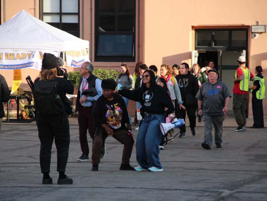 People gather outside a building; some pose for a photo while others walk or stand nearby. A white tent and workers in safety vests are visible in the background.
