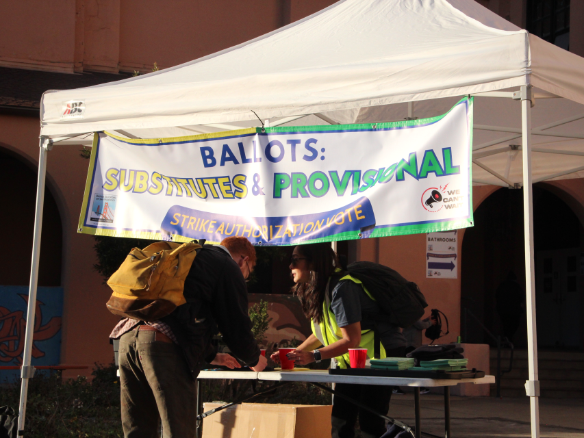 Two people stand at a table under a canopy with a sign reading "Ballots: Substitutes & Provisional, Strike Authorization Vote" outside a building.