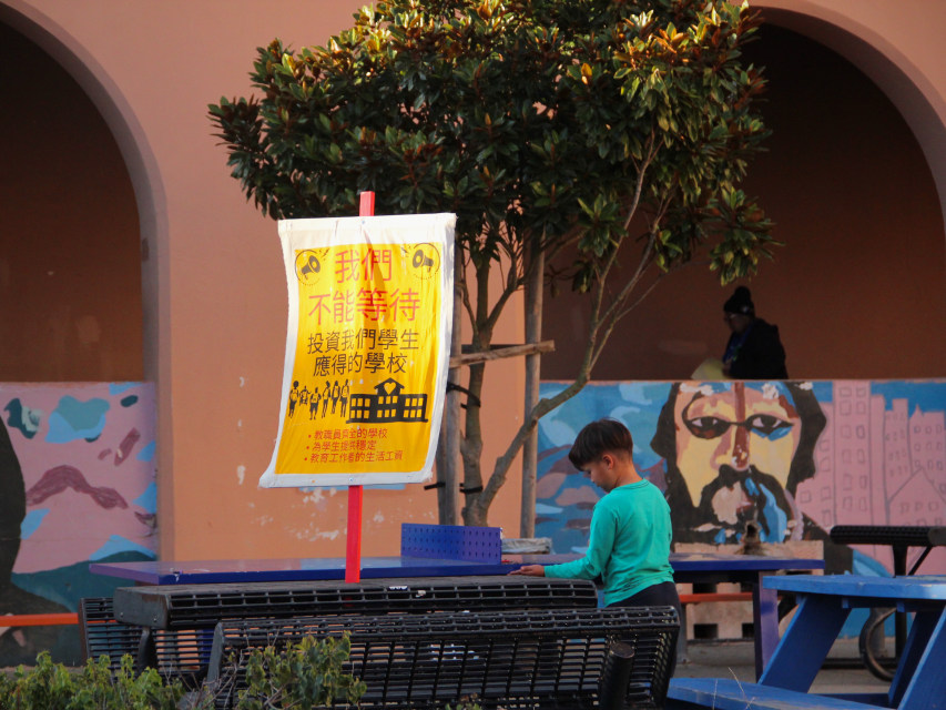 A child stands near a bench in front of a sign with Chinese text and a mural depicting a face on a wall in the background.