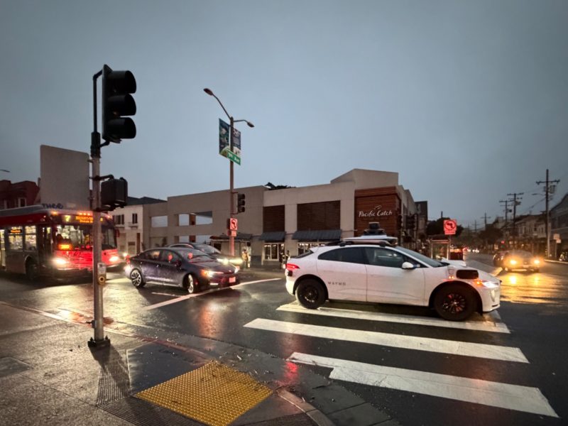 Several vehicles, including a bus and a white SUV, wait at an intersection on a wet, overcast evening in a commercial urban area.