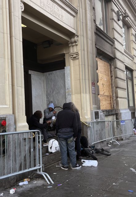 Several people gather on the steps of a building entrance, surrounded by metal barricades and scattered belongings on a littered sidewalk.