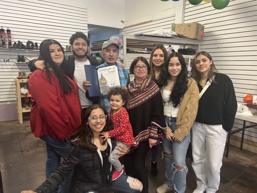 A group of nine people, including a toddler, pose and smile inside a store. One person holds a certificate; shelves and boxes are visible in the background.