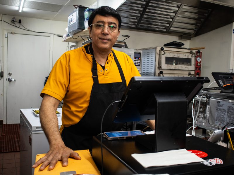 A man wearing glasses, a yellow shirt, and a black apron stands behind a counter in a commercial kitchen, with a cash register and electronic equipment nearby.
