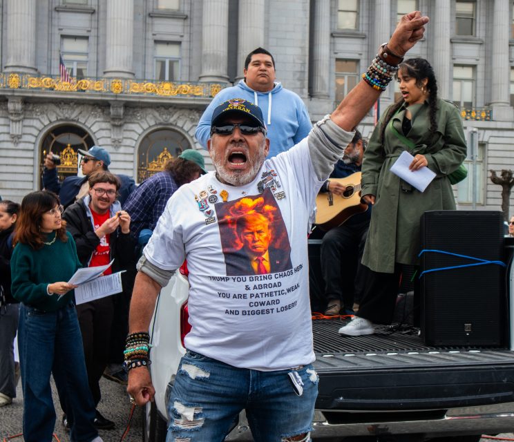 A man raises his fist and shouts at a rally in front of a government building, wearing a t-shirt with a graphic and text, surrounded by other people, some holding papers and a guitar.