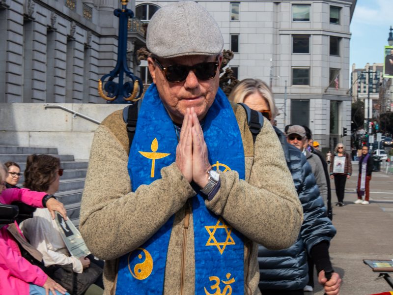 A man wearing sunglasses, a flat cap, and a blue stole with religious symbols stands outdoors with hands pressed together in prayer.