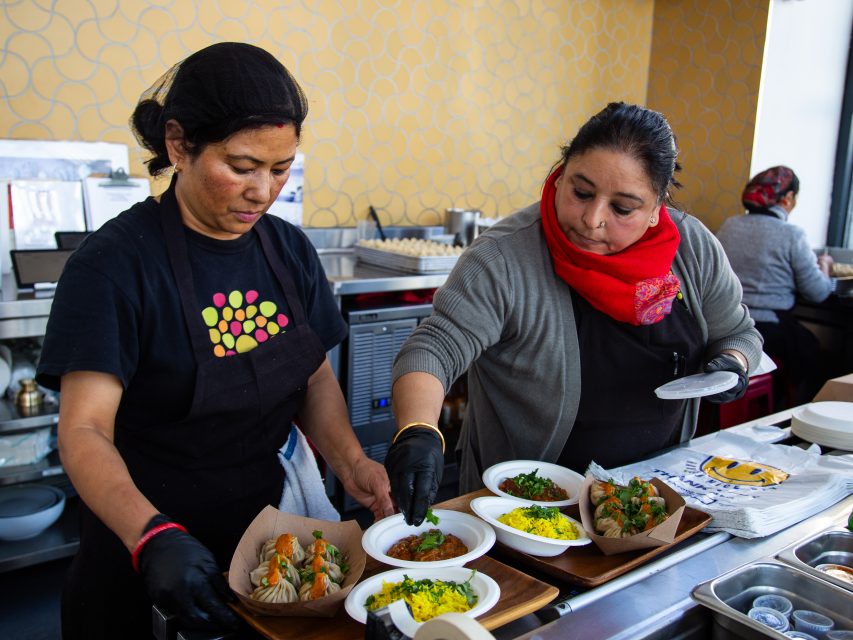 Two women prepare and arrange plated dishes with food in a kitchen, wearing gloves and working together at a counter.
