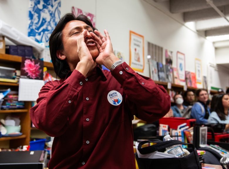 A person in a maroon shirt with a "FAITH" sticker is shouting or calling out in a busy room with people and various supplies in the background.