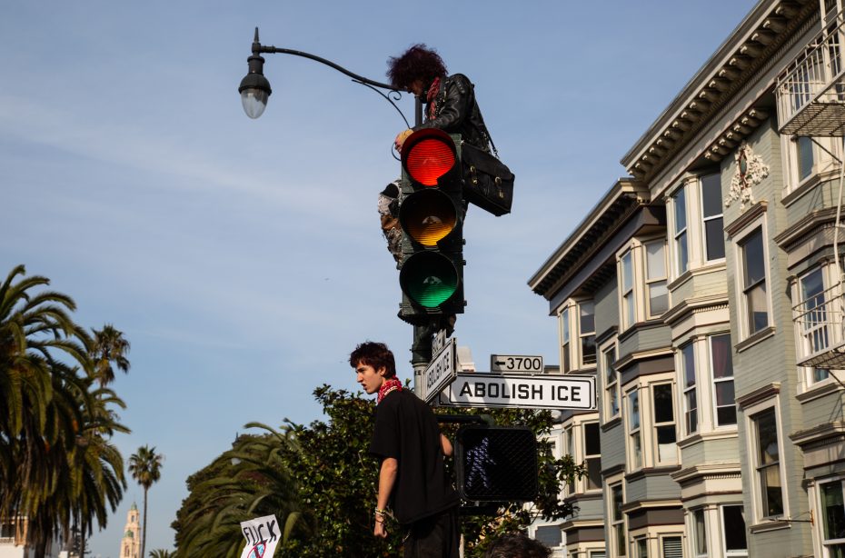 Two people at a traffic light; one sits atop the signal while another stands below. Signs on the pole read "ABOLISH ICE." Residential buildings and palm trees are in the background.