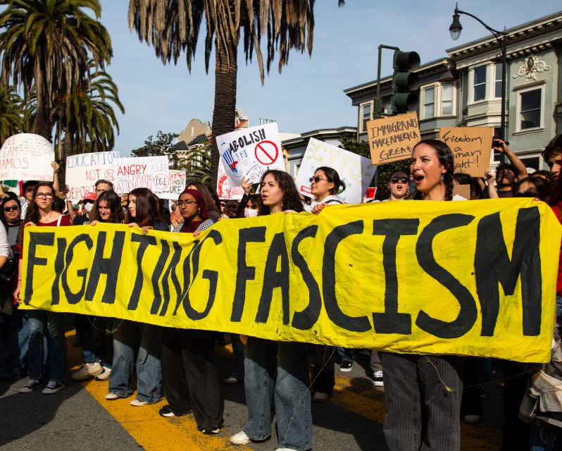 A group of protesters hold a large yellow banner reading “FIGHTING FASCISM” at a demonstration, with various signs visible in the background.