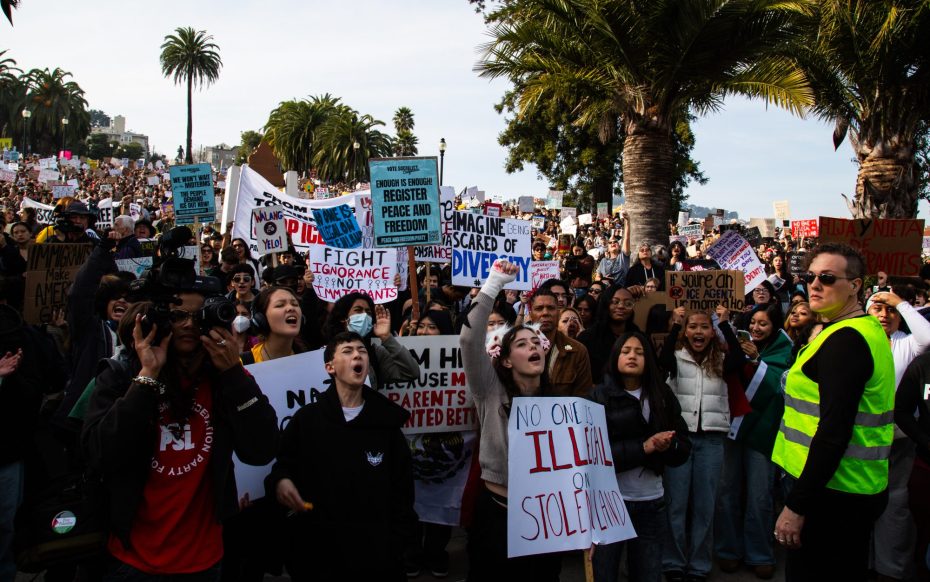 A large crowd holds signs and protests outdoors in a palm-lined area; a person in a yellow vest stands at the front.
