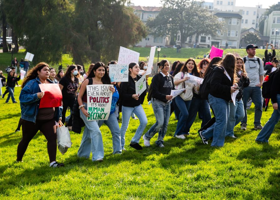 A group of people march on a grassy area holding protest signs, including one that reads, "This isn't even about politics. It's about human decency.
